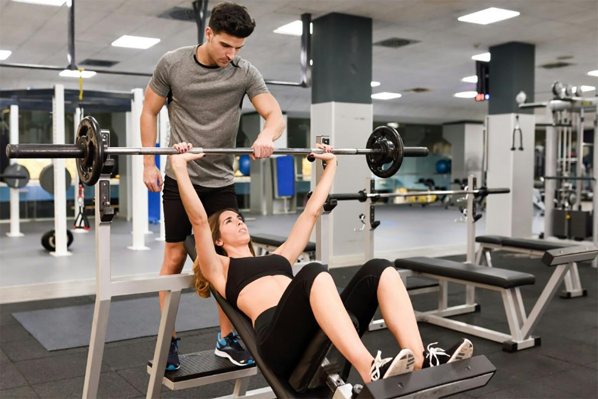 A woman undergoing physical therapy bench presses a barbell on a decline bench while a man spotting her ensures safety in a gym setting.